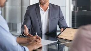 Un asesor financiero sonriente en traje gris muestra gráficos de rendimiento en una tablet durante una reunión de negocios con clientes.