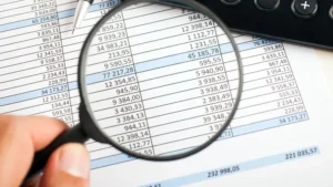 Mano sosteniendo una lupa sobre una hoja de balance financiero impresa con columnas de números y cifras.