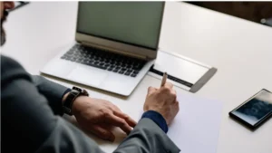 Primer plano de un profesional en traje escribiendo sobre documentos de planificación fiscal con un bolígrafo de lujo, junto a una laptop en un escritorio.