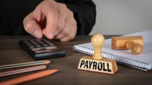 Sello de madera con la palabra Payroll junto a las manos de un contador utilizando una calculadora sobre un escritorio.